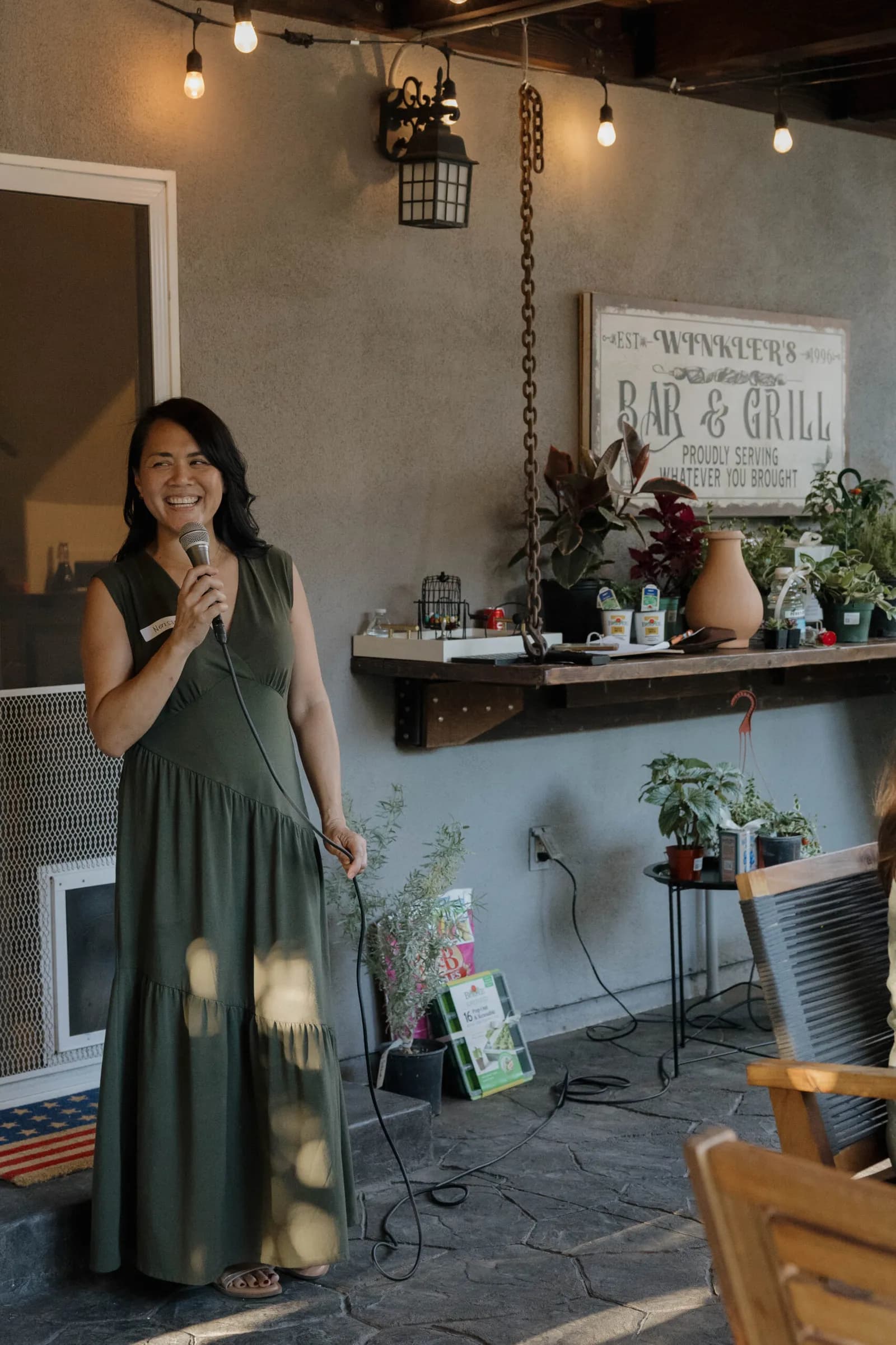 Nerissa hosting a bingo night on a backyard patio, microphone in hand, plant prizes on the shelf behind her
